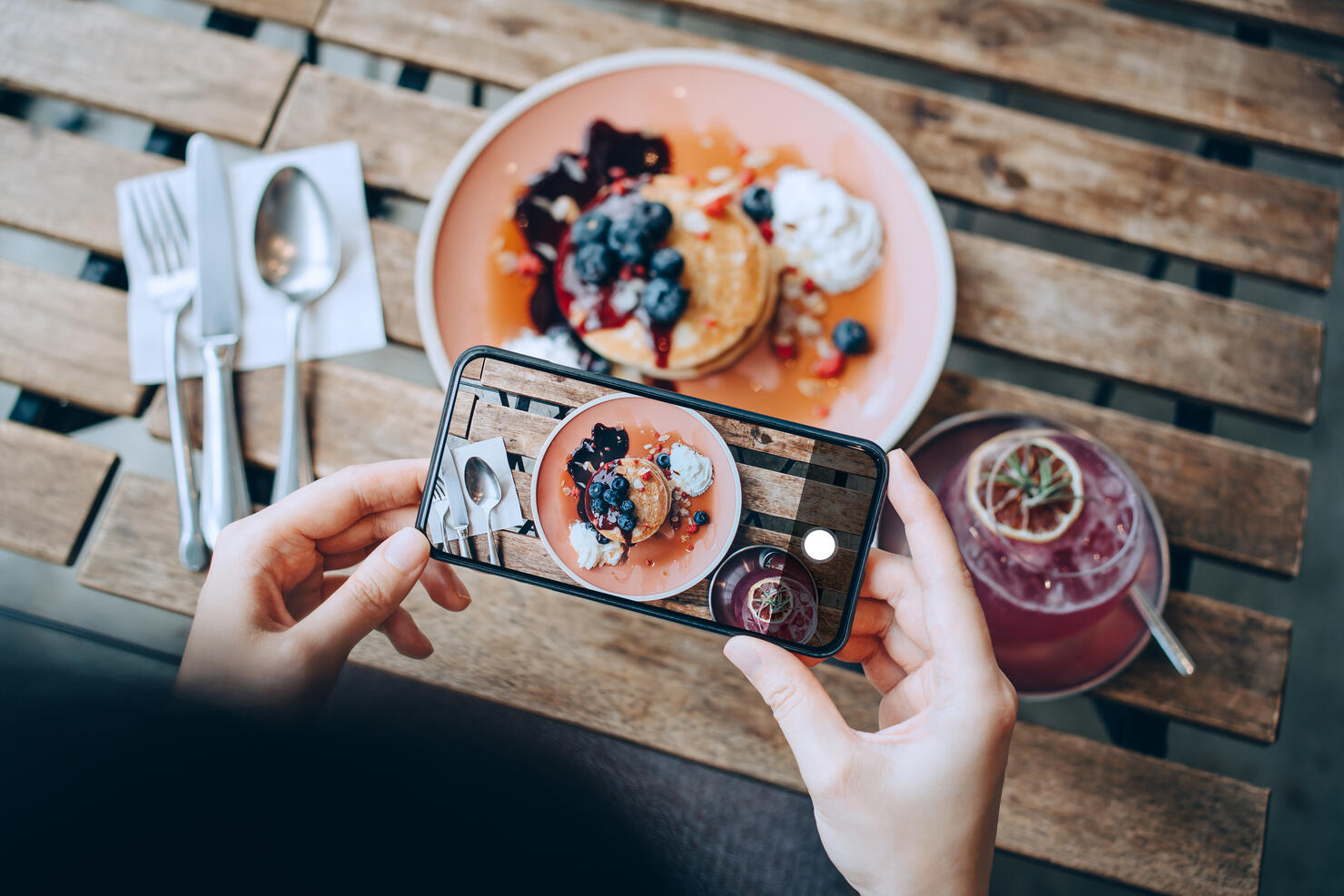 Overhead view of young Asian woman taking photos of delicious freshly served pancakes with blueberries and pink lemonade with smartphone before eating it in cafe. Eating out lifestyle. Camera eats first culture