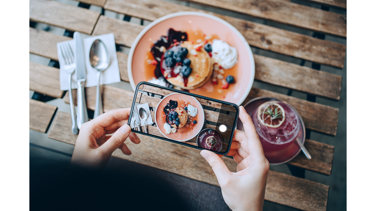 Overhead view of young Asian woman taking photos of delicious freshly served pancakes with blueberries and pink lemonade with smartphone before eating it in cafe. Eating out lifestyle. Camera eats first culture