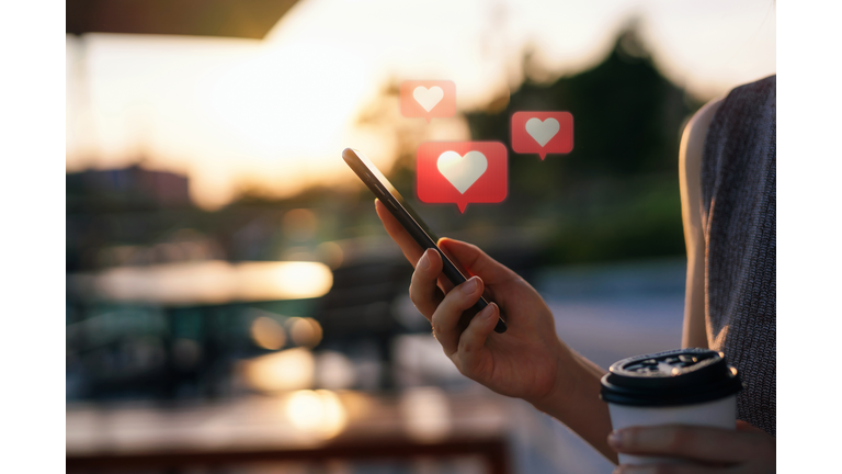 Close up of young woman checking social media network on smartphone on the go in the city. Mobile phone with heart emoji speech bubble message on the screen. People engaging in networking with technology. Social media addiction concept