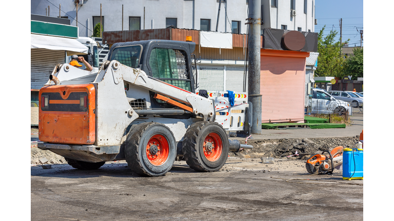 A highly manoeuvrable construction vehicle with a jackhammer attached is parked at a work site with a city street being repaired in the background.