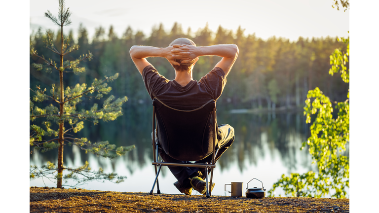 Man is sitting in a camping chair on the background of a forest lake on a beautiful summer evening.