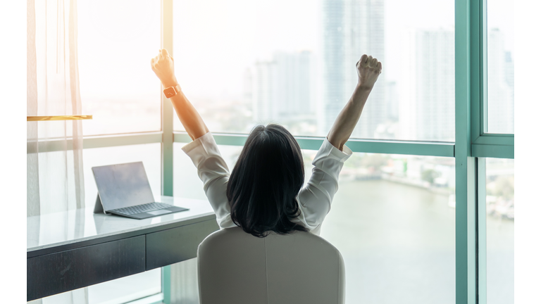 Business achievement concept with happy businesswoman relaxing in office or hotel room, resting and raising fists with ambition looking forward to city building urban scene through glass window