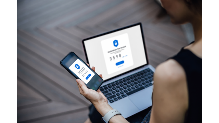 Over the shoulder view of young woman using mobile device with Two-Factor Authentication (2FA) security while logging in securely to her laptop. Privacy protection, internet and mobile security