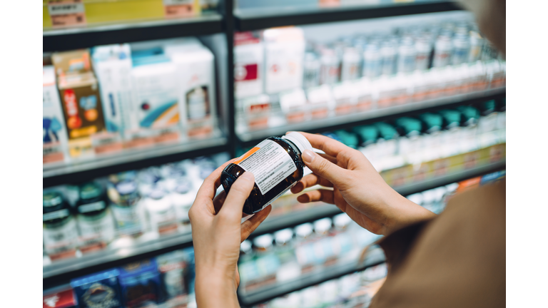 Over the shoulder view of young Asian woman browsing through medical products and reading the label on a bottle of medicine in front of the shelves in a pharmacy