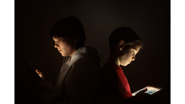 Brother and sister use smartphone and digital tablet while sitting back to back at night at home