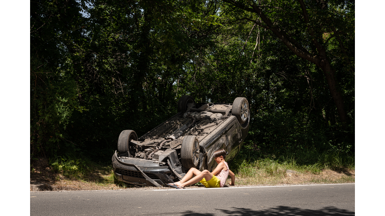 A boy in shorts and a T-shirt, a participant in a car accident, is lying in shock next to an overturned car by the roadside.