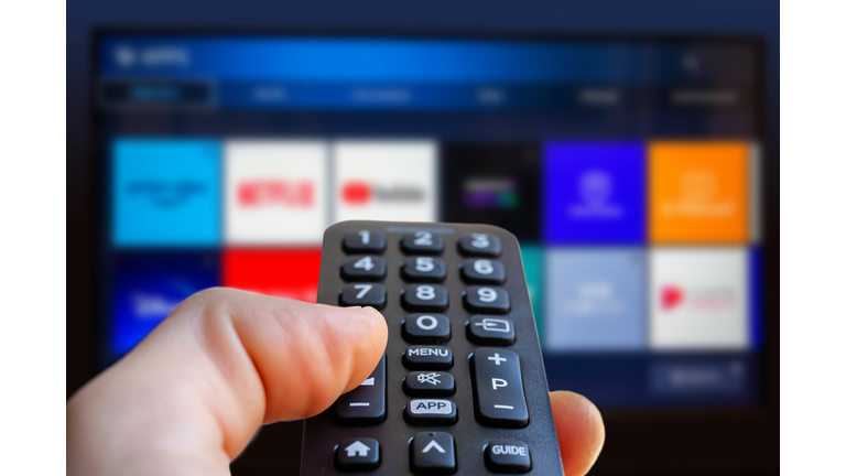 A man is holding a remote control of a smart TV in his hand. In the background you can see the television screen with streaming entertainment apps for video on demand