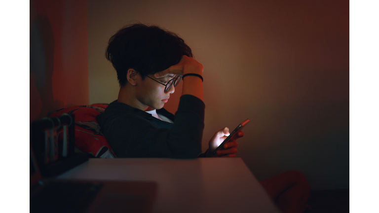 The upset teenage Asian boy is reading alone on his smartphone late at night, caught in the grips of adolescent social media addiction