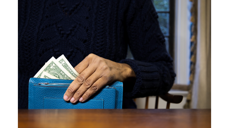 black woman holding vibrant blue wallet with multiple $1 u.s. paper bills showing while sitting at desk at home