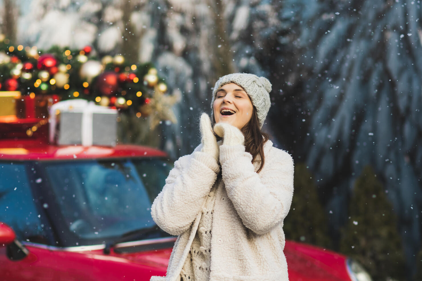 Positive woman in wool mittens and fur jacket standing near car with green christmas tree and presents on top.
