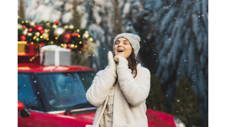 Positive woman in wool mittens and fur jacket standing near car with green christmas tree and presents on top.