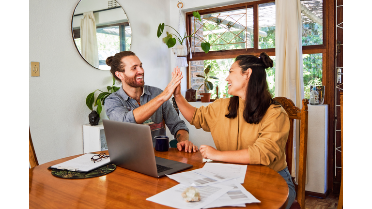 Shot of a happy young couple sharing a high five while using a laptop and checking paperwork at home