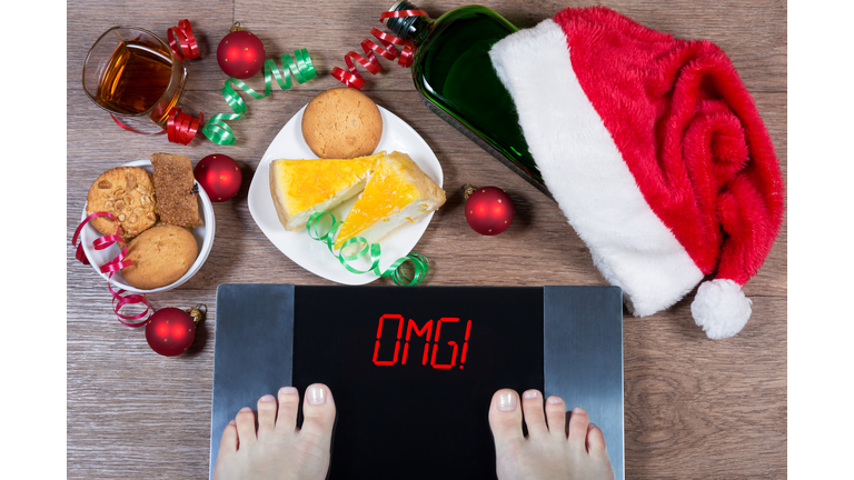 Female feet on digital scales with sign "omg!" surrounded by Christmas decorations, bottle, glass of alcohol and sweets. Consequences of overeating and unhealthy lifestile during holidays.