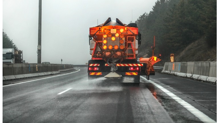 Orange highway maintenance gritter truck spreading de-icing salt, crystals dropping on the ice covered asphalt road during overcast day