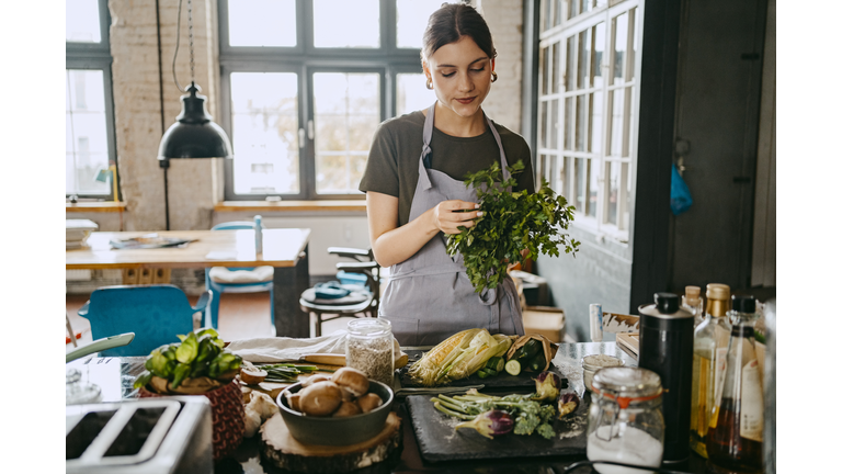 Female chef wearing apron doing quality check of cilantro standing in studio kitchen