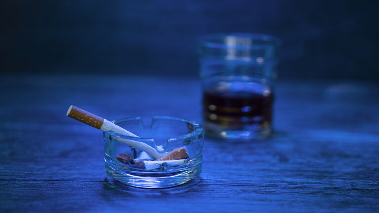 View of an ashtray filled with cigarettes. The focus is on the ashtray. There is also a glass of alcoholic beverage on in the back. There is a blue light. They are on a wooden table.