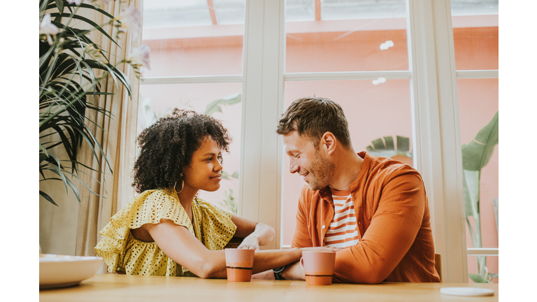 A beautiful young couple looks very much in love as they sit at a dining table sipping hot drinks and flirt.