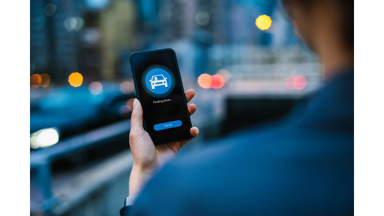 Over the shoulder view of young Asian woman arranging a taxi service with mobile app on smartphone in city street in the evening, with illuminated city scene in background. Lifestyle and transportation technology. Convenience and trustworthy car service