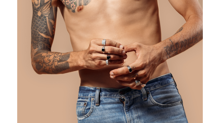 Freestyle. Man with bare upper body standing isolated on beige touching fingers wearing rings