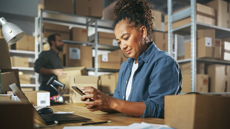 Inventory Manager Using Smartphone to Scan a Barcode on Parcel, Preparing a Small Cardboard Box for Postage. Black Multiethnic Small Business Owner Working on Laptop in Warehouse.