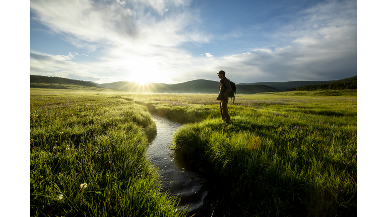 A man standing next to a small stream flowing through a beautiful green field at sunrise.