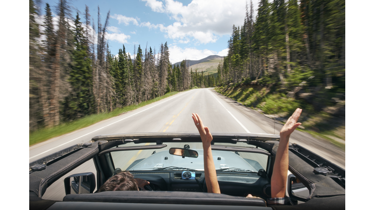 Road trip couple driving convertible on rural highway with hands raised, Breckenridge, Colorado, USA
