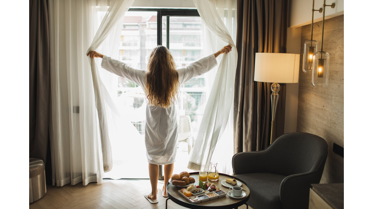 View from behind of woman opening curtains in hotel room. Morning awakening