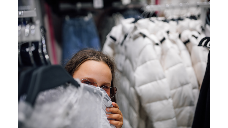 Playful young girl hides behind tack with winter jackets enjoying fun games in favorite clothing store