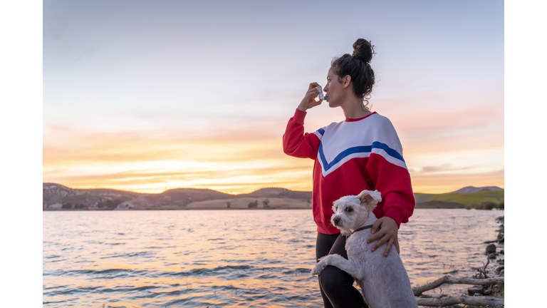 Millennial Woman Using Her Asthma Inhaler At The Lake Shore At Sunset