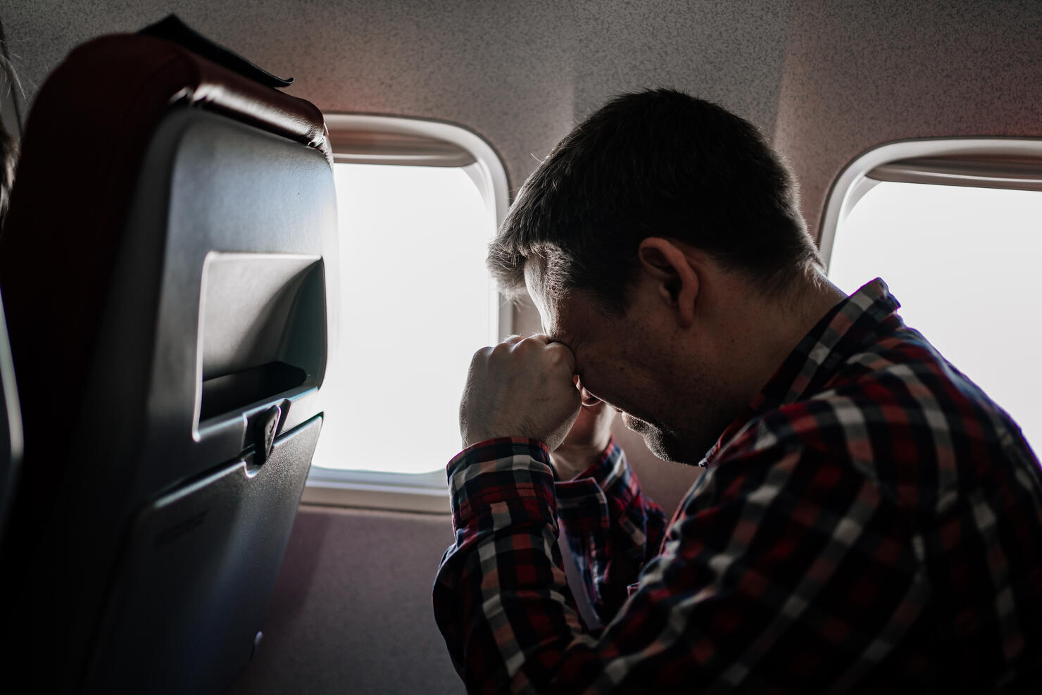 man in plaid shirt rubs eyes with his hands in seat of passenger on plane.