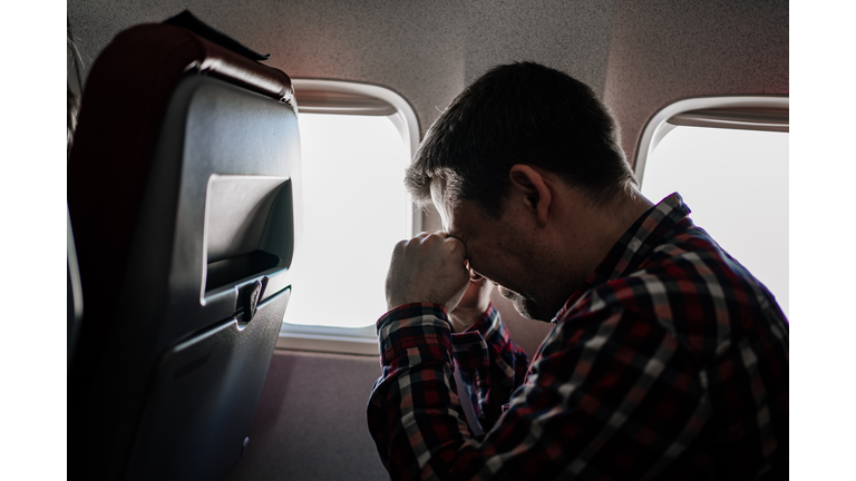 man in plaid shirt rubs eyes with his hands in seat of passenger on plane.