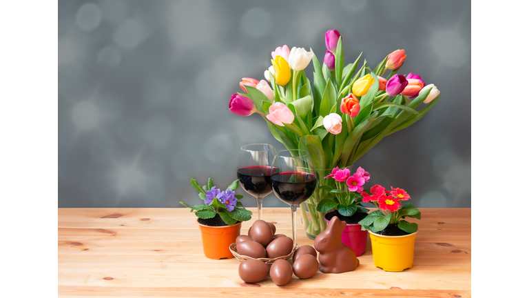 Easter concept. Colorful tulips in vase, chocolate bunny, glasses of red wine, primula flowers and chocolate Easter eggs in wicker basket on wooden table.