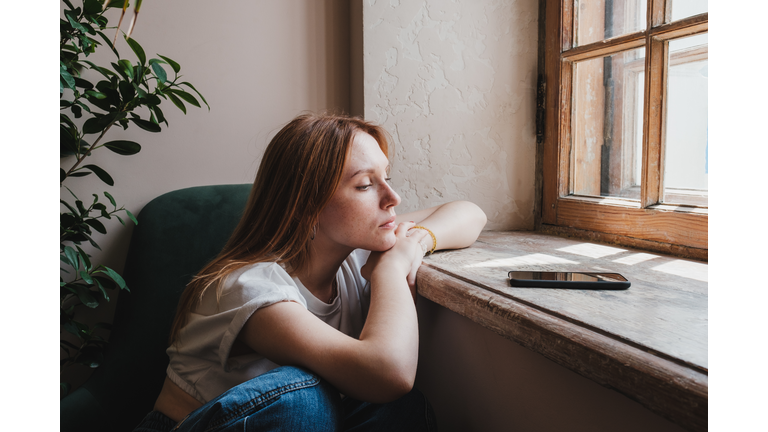 Upset redhead teen girl sitting by window looking at phone waiting call or message