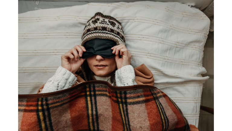 A young woman in a warm hat and sweater lifts a sleeping mask. The girl is covered with a plaid blanket and lies in bed. Close-up. Scandinavian style.