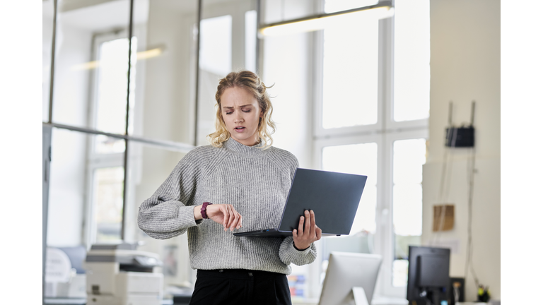 Young woman standing in office with laptop checking the time