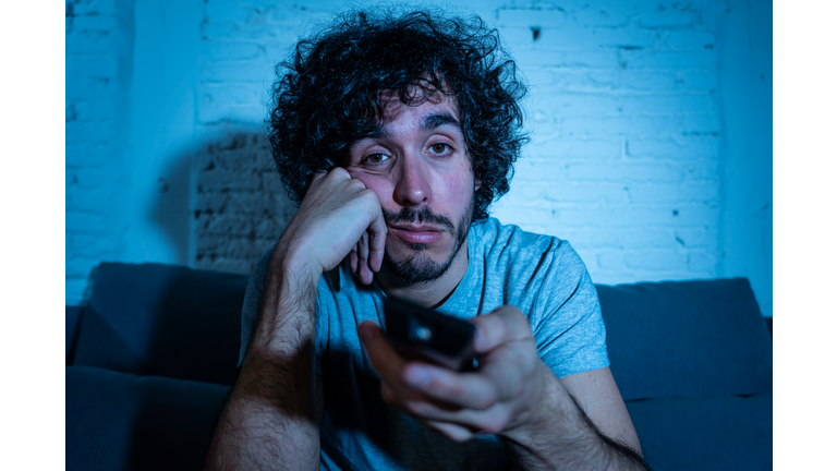 Young bored man on couch using TV remote control zapping for another movie or show late at night. Looking disinterested and sleepless. In entertainment People insomnia and Sedentary lifestyle concept.