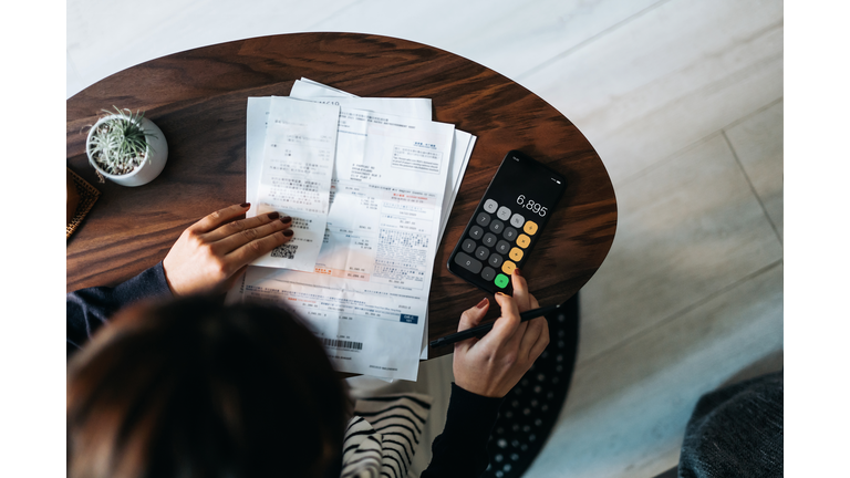 Overhead view of young Asian woman managing personal banking and finance at home. Planning budget and calculating expenses while checking her bills with calculator. Managing taxes and financial bills. Home budgeting. Concept of finance and economy