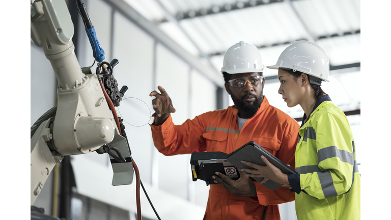 Intelligent Design of Robotic Welding Process Parameters in Manufacturing Industry. A Female Manufacturing Process Engineer and a welder is discussion in-front of a robotics welding in production line about welding process parameter to improve auto part.