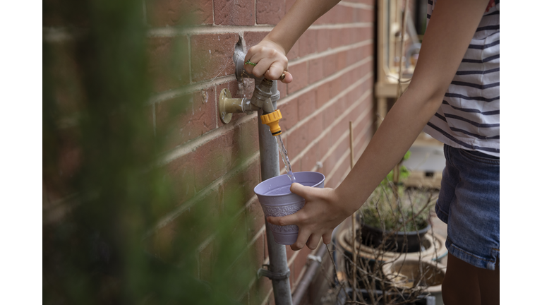 Child filling a decorative metal pot with water in a back yard from an outdoor tap in summertime.