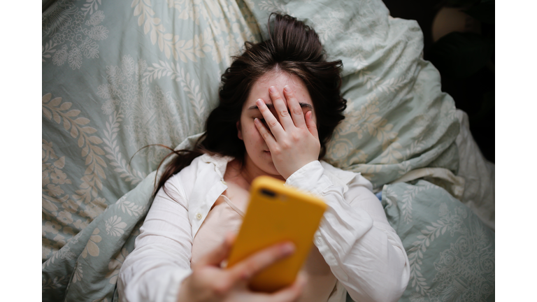 Sleepy overweight young woman with flowing hair using phone on bed, close face palm. Top view