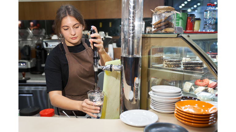 A young beautiful woman in a brown apron is standing and pouring water into a glass. The concept of a small business. Work for yourself.