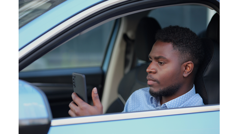 African American driver with phone sitting at car steering wheel