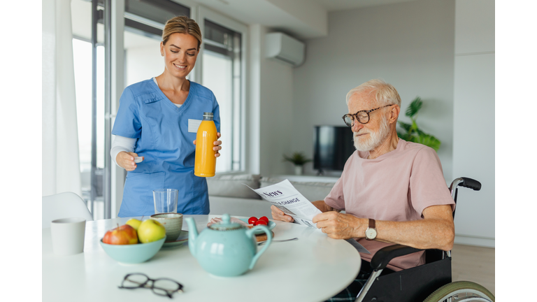 Caregiver preparing breakfast for senior man in the morning, pouring orange juice in glass. Elderly man in wheelchair at home need help from nurse.