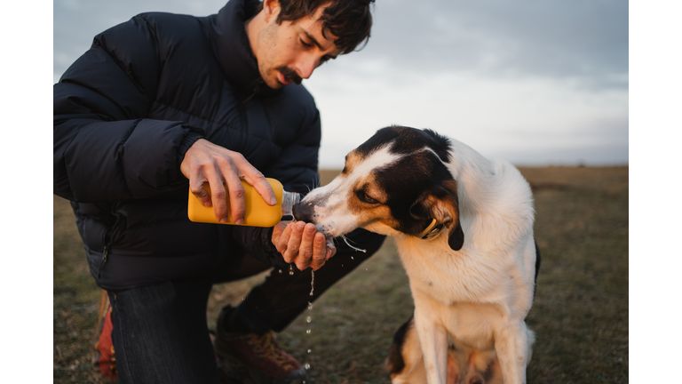 young white man giving his dog drinking water during sunset