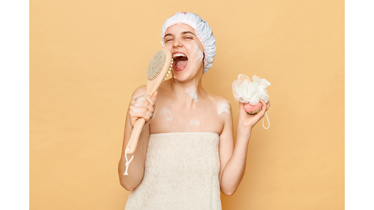 Extremely happy woman wearing bath cap and wrapped in towel holding sponge in hands and wooden body brush like microphone singing with positive emotions in morning.