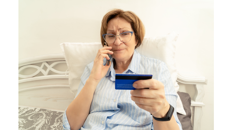 Elderly doubtful woman talking on a mobile phone and holding a bank card in her hand