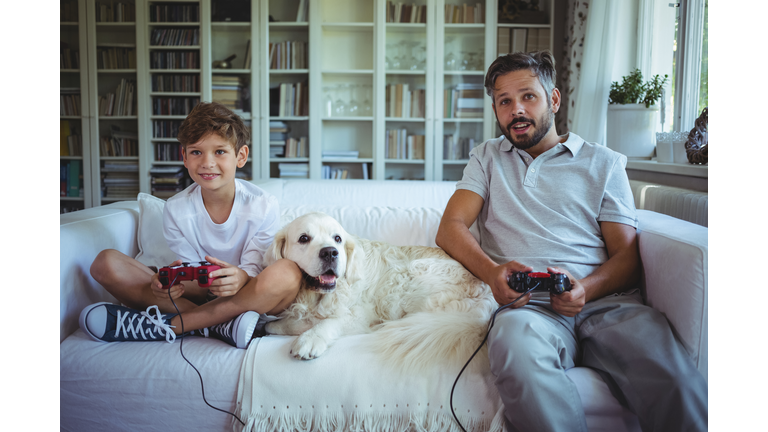 Father and son sitting on sofa with pet dog and playing video games