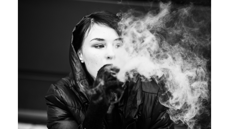 Vape teenager. Young pretty white girl in the hood smoking an electronic cigarette opposite modern background on the street in the winter. Bad habit. Black and white.