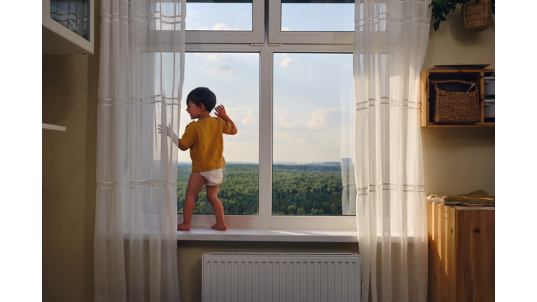 The child is alone by the window, lost in thought and risking their safety.