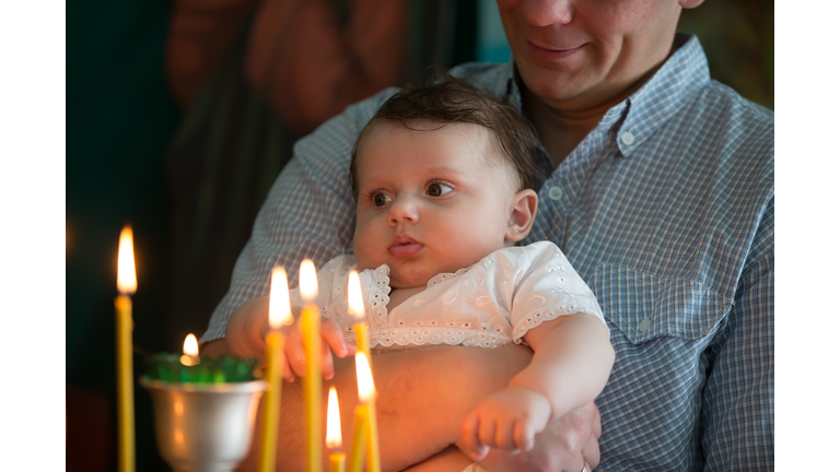 The father's hands hold the baby in his arms in the church. The kid looks at the candles burning in the temple. Child in church